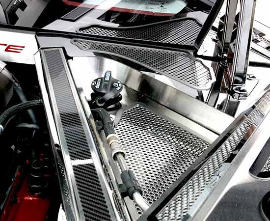 Close view of the perforated stainless rear cowl cover beside a C8 Corvette Coupe engine-hatch hinge, with surrounding trim shown
